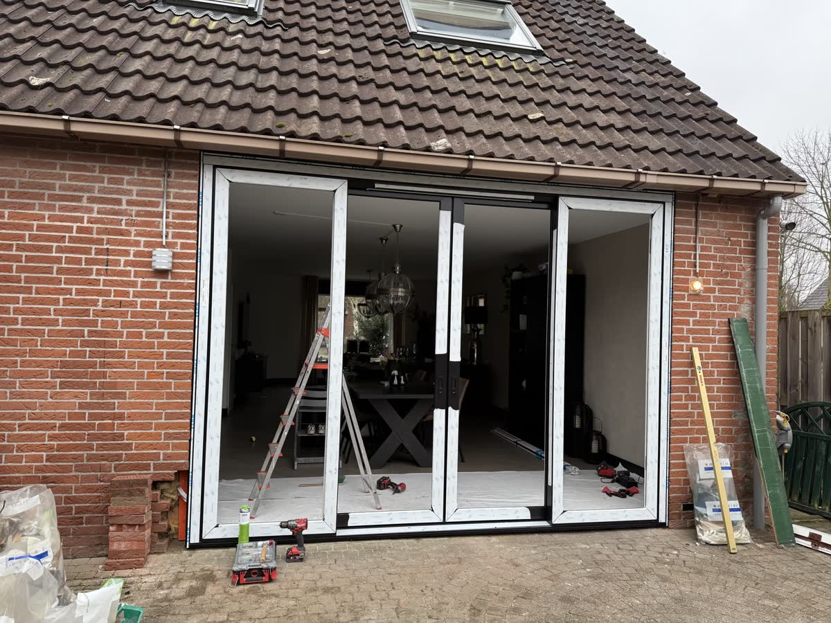 Aanbouw van rode baksteen tijdens de montage: het nieuwe kozijn staat al in de opening, met zicht in de woonkamer