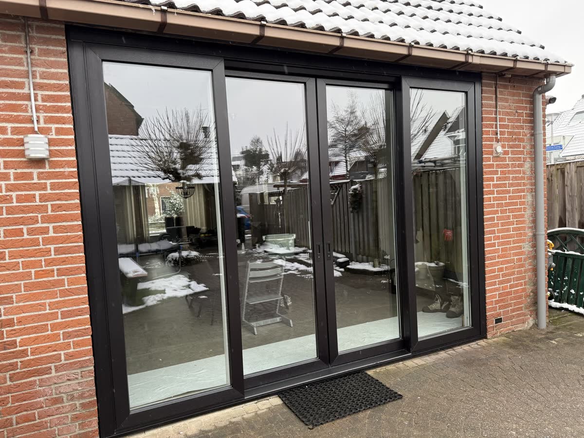 Donkere kunststof pui met vier grote glasvlakken in een aanbouw van rode baksteen