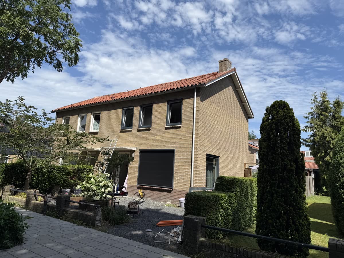 Woning van gele baksteen met nieuwe kunststof ramen en een rolluik, aan een tuin vol groen