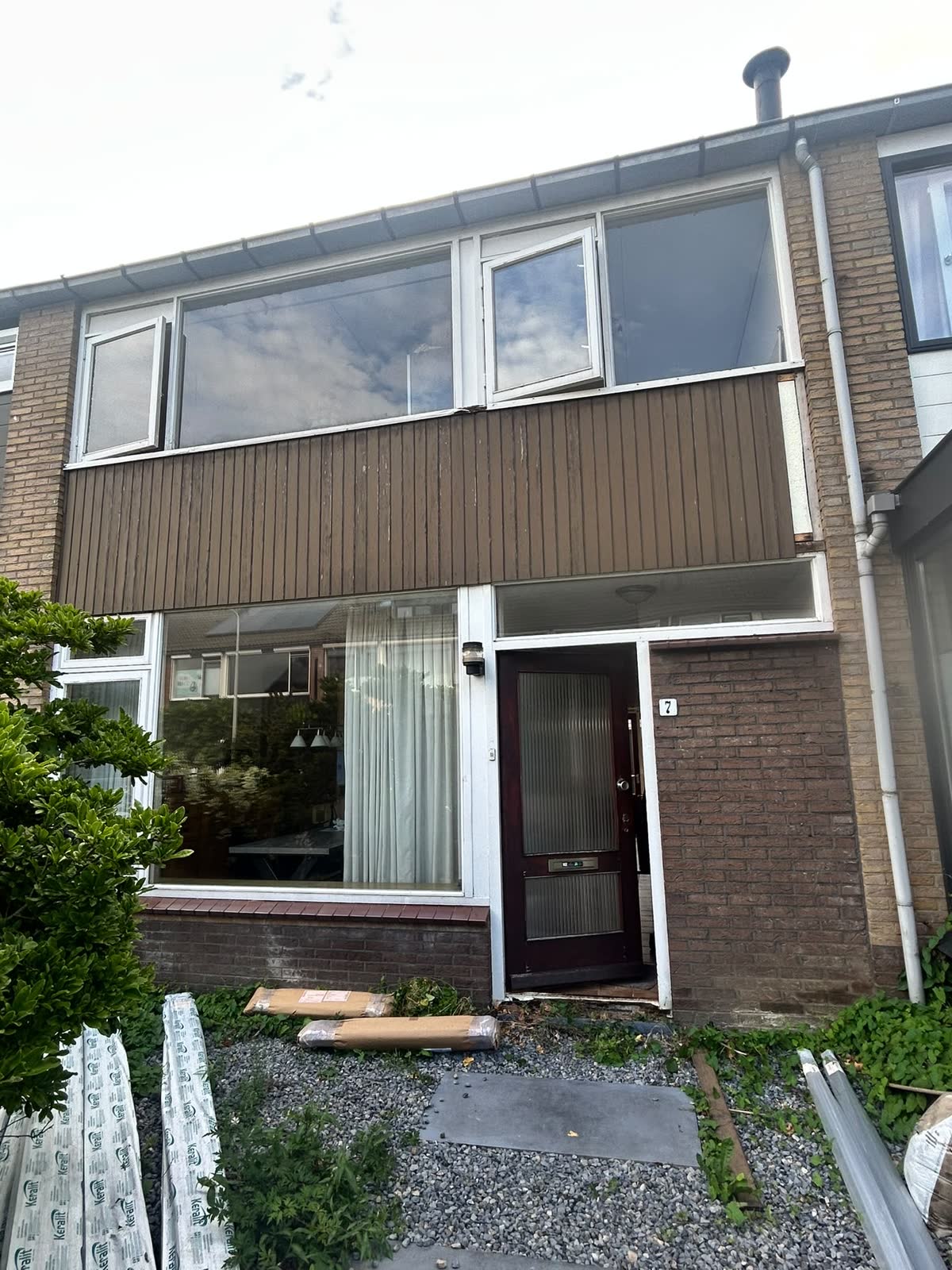 Dezelfde voorgevel voor de vervanging, met witte kozijnen en een bruine gevelbekleding