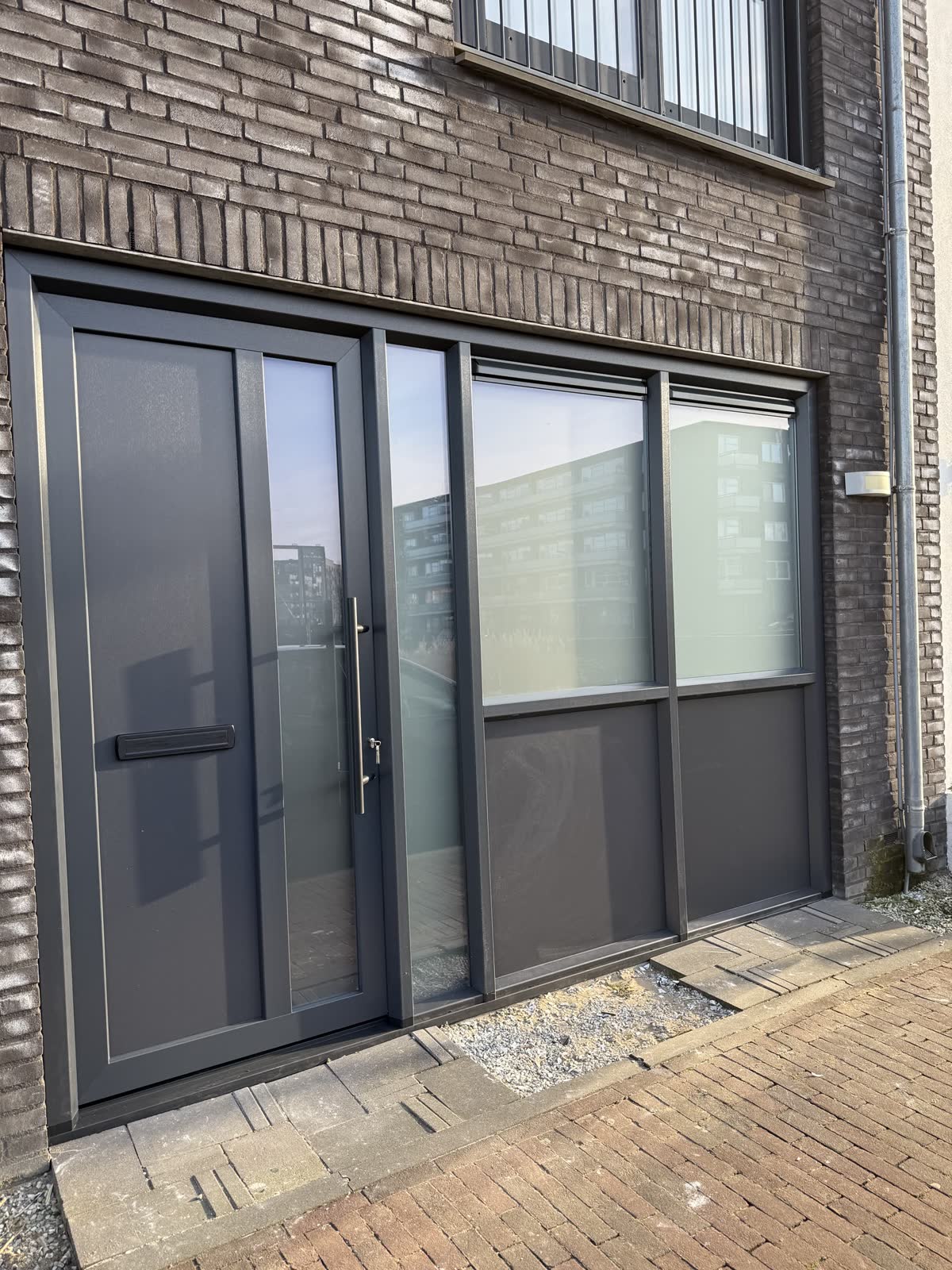 Antraciet kunststof deur met een glazen zijlicht en vaste glasvlakken ernaast, in een donkere bakstenen gevel
